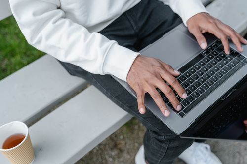 Free Man working on a laptop outdoors with a coffee cup beside him, showcasing digital nomad lifestyle. Stock Photo
