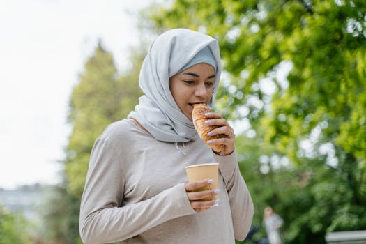 A young woman in a hijab enjoying a croissant and drink outdoors in a spring setting.
