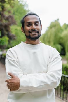 Portrait of a smiling man standing with arms crossed in a lush green park on a clear day.