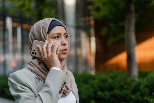 A professional Muslim woman in hijab talks on her smartphone while standing outdoors in business attire.