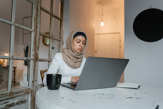 Muslim woman in hijab typing on a laptop indoors with a coffee mug nearby at a table.