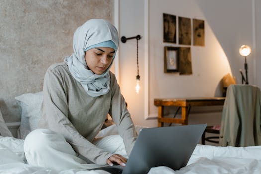 A young Muslim woman wearing a hijab works on her laptop in a stylish, contemporary bedroom setting.