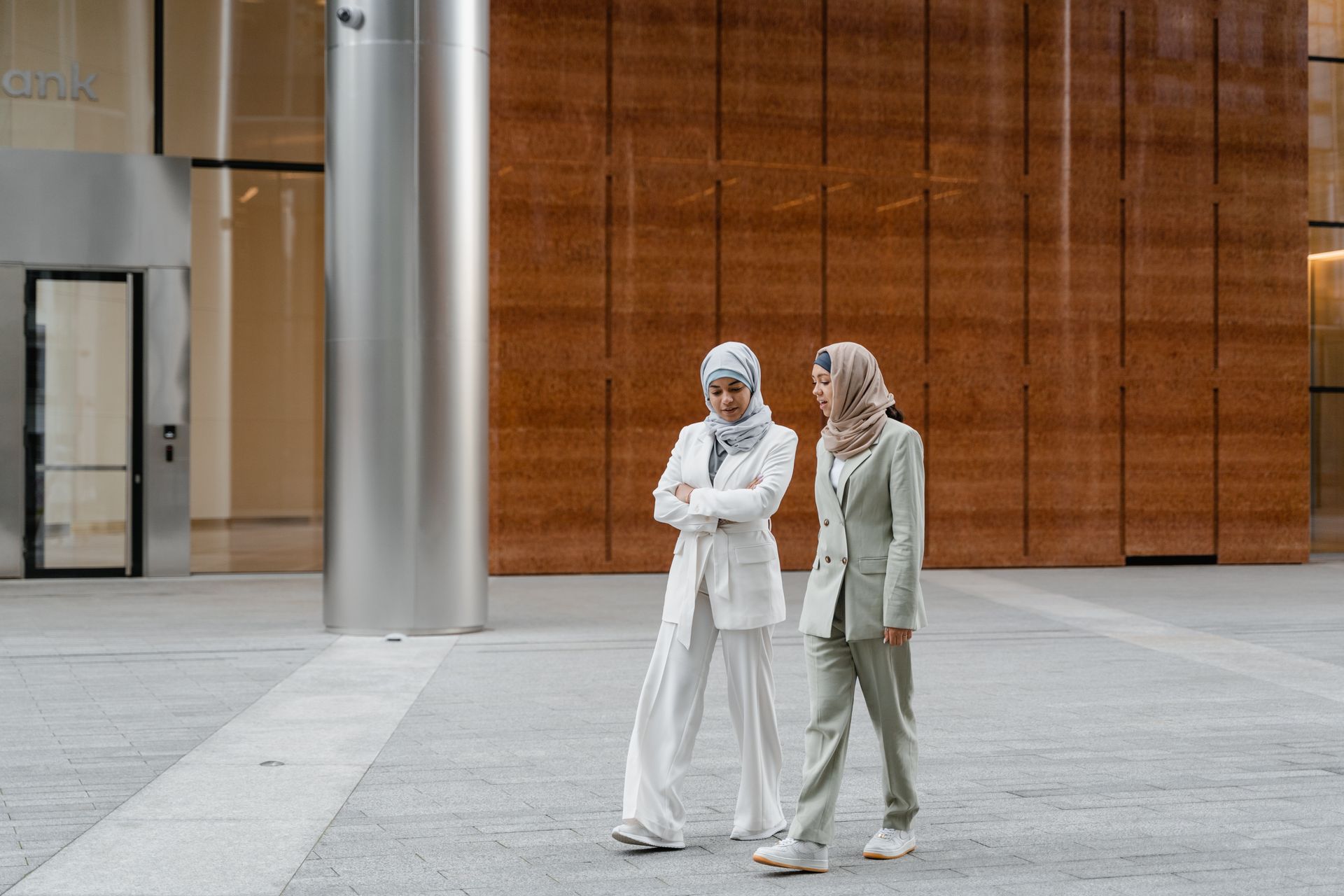 Two Muslim women walking in business attire outside a modern building, discussing work.