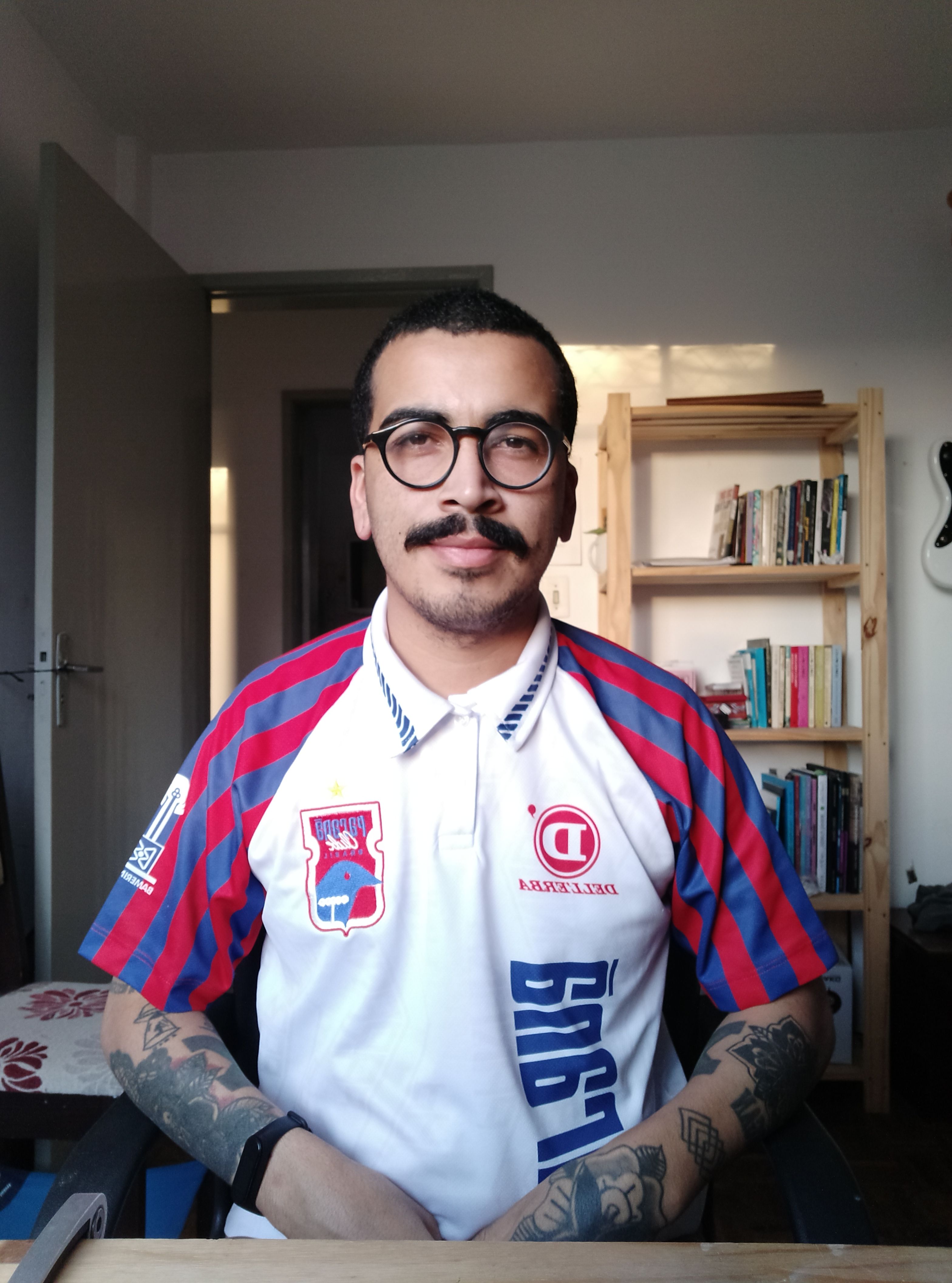 Portrait of a man indoors with tattoos, eyeglasses, and a mustache wearing a sports polo shirt