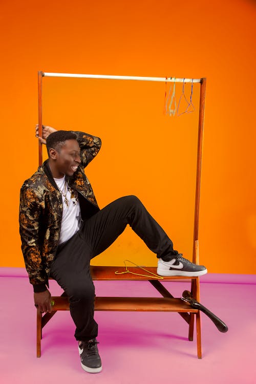 Full body of positive African American male in trendy outfit sitting on empty clothing rack with hangers on orange background in studio