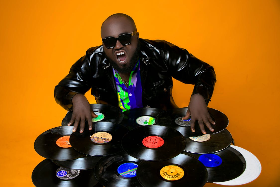 Skilled African American male DJ in leather jacket standing in studio while scratching abundance of various vinyl records on orange background