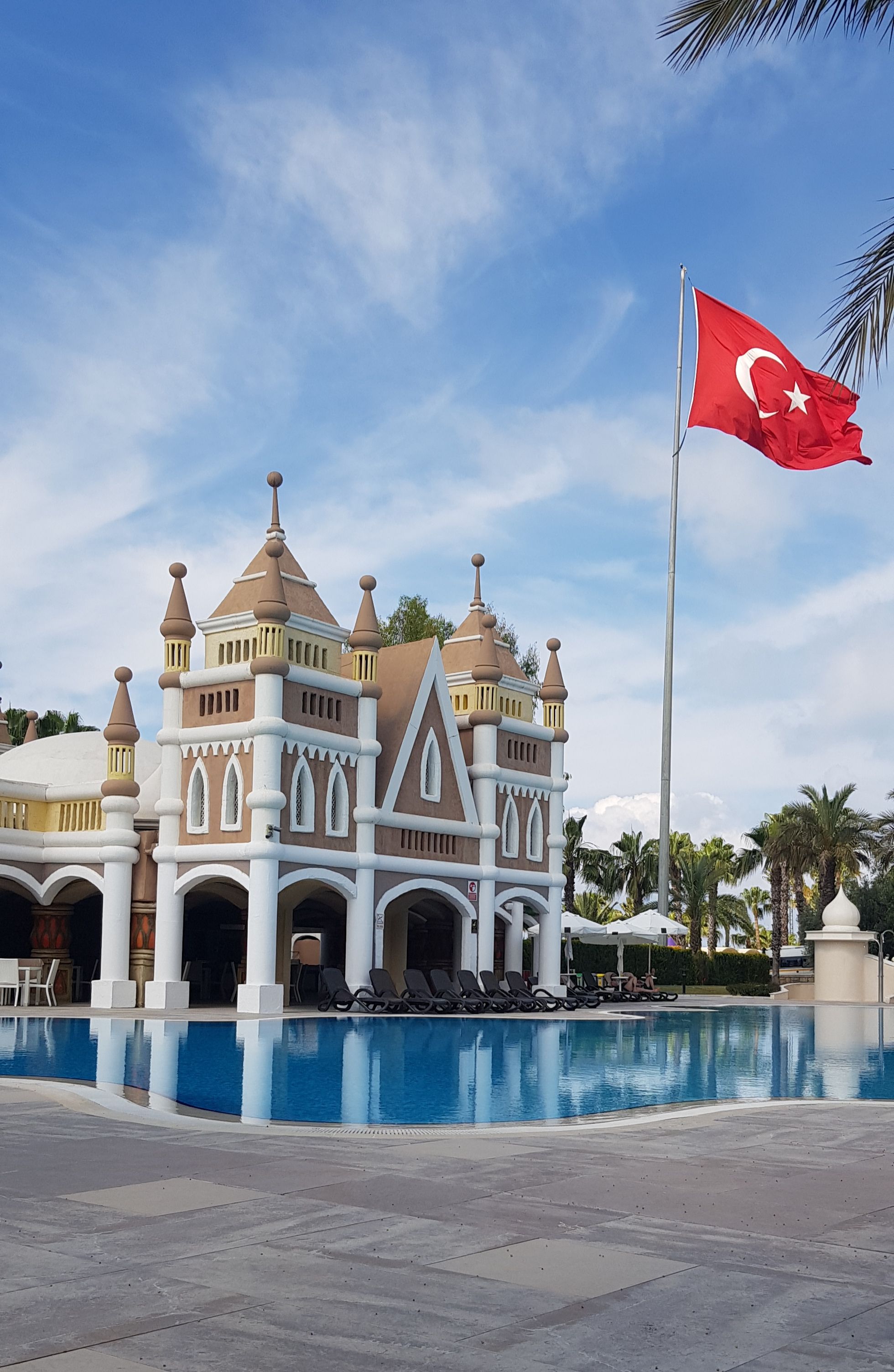 Elegant hotel exterior with a Turkish flag and a swimming pool, under a blue sky.