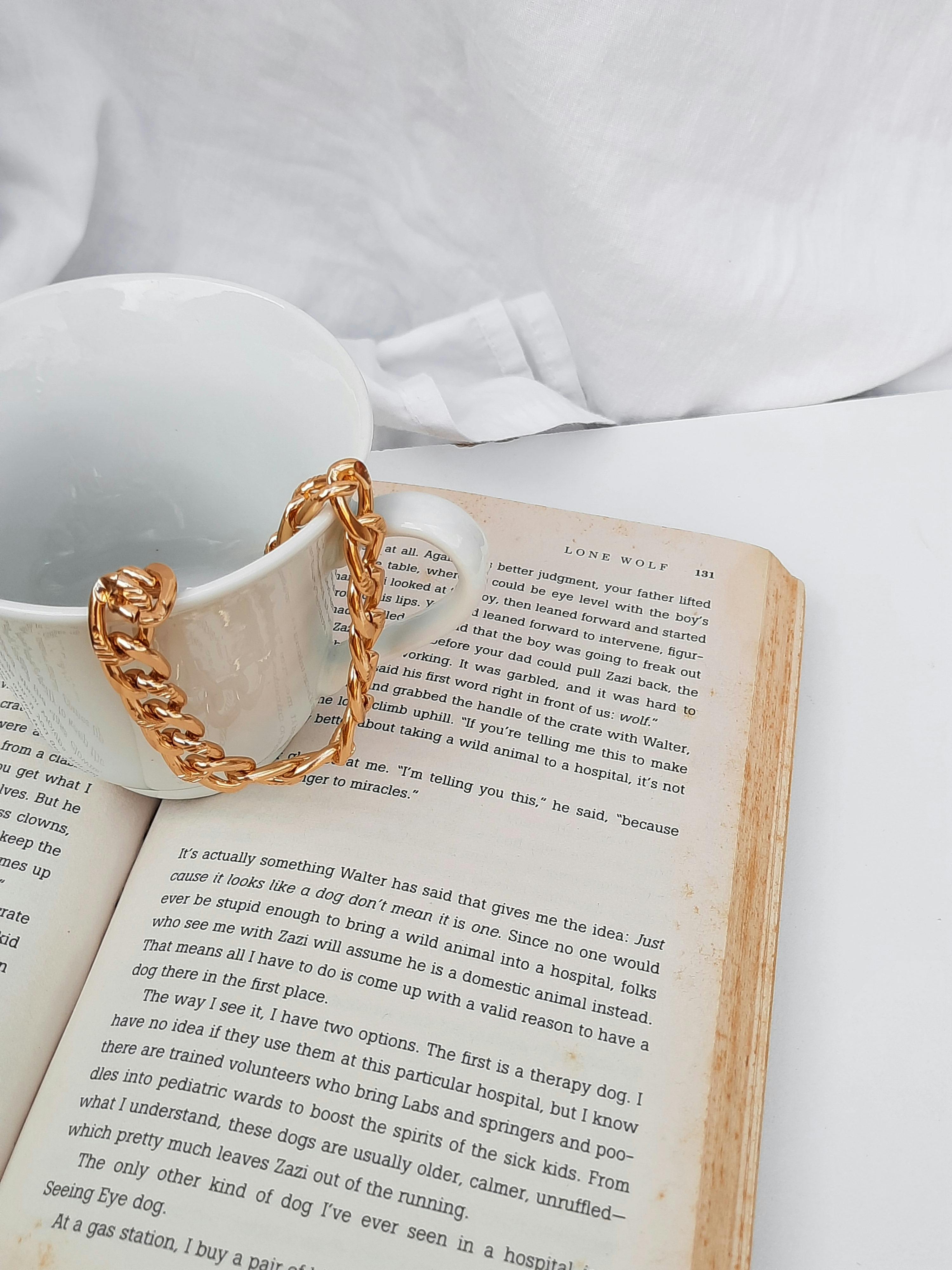 Free A stylish still life featuring a ceramic mug adorned with a gold chain, resting on an open book. Stock Photo