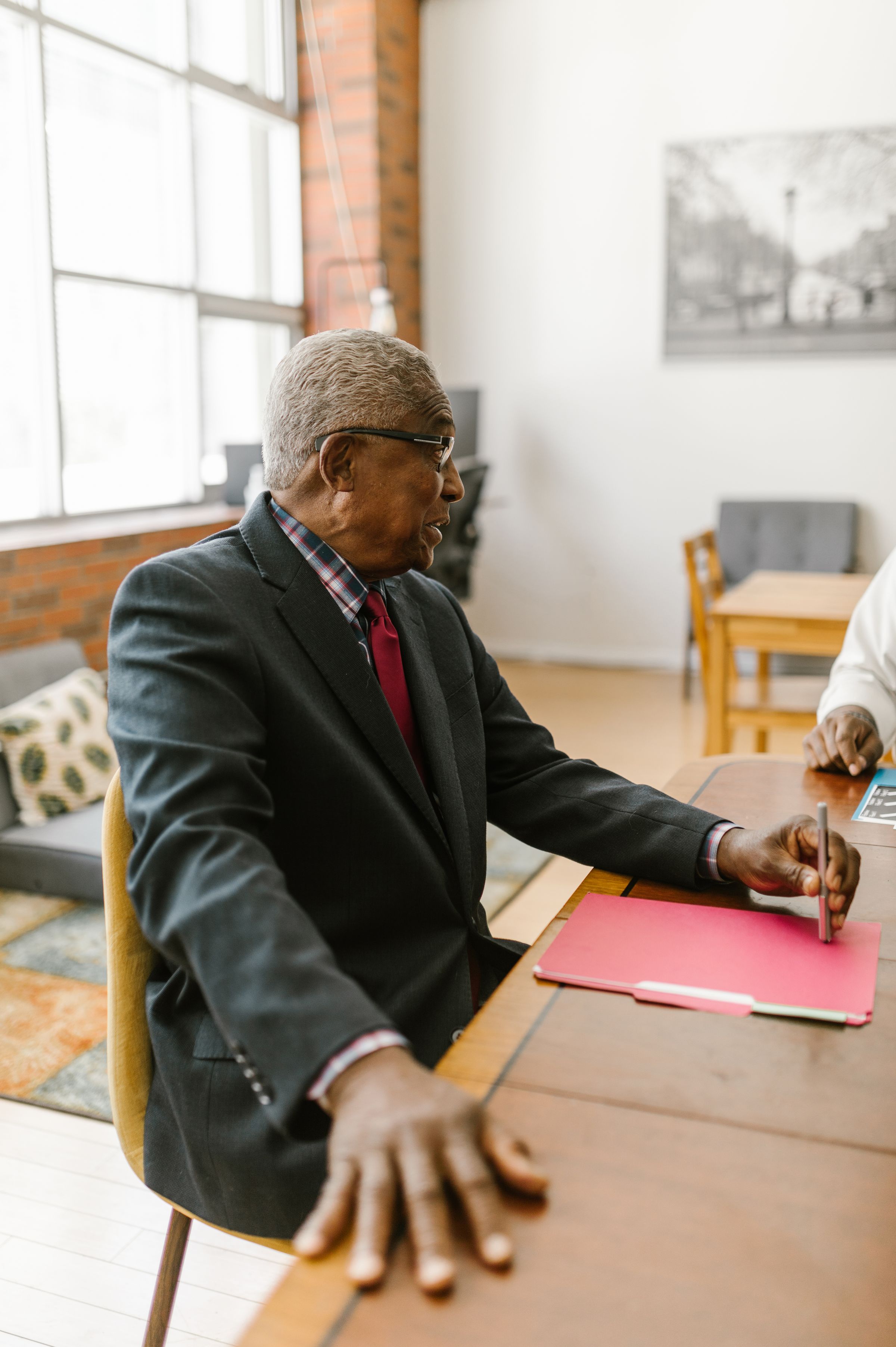 Senior man in a dress shirt and suit jacket holding a pen over a folder at an interview table in a bright office