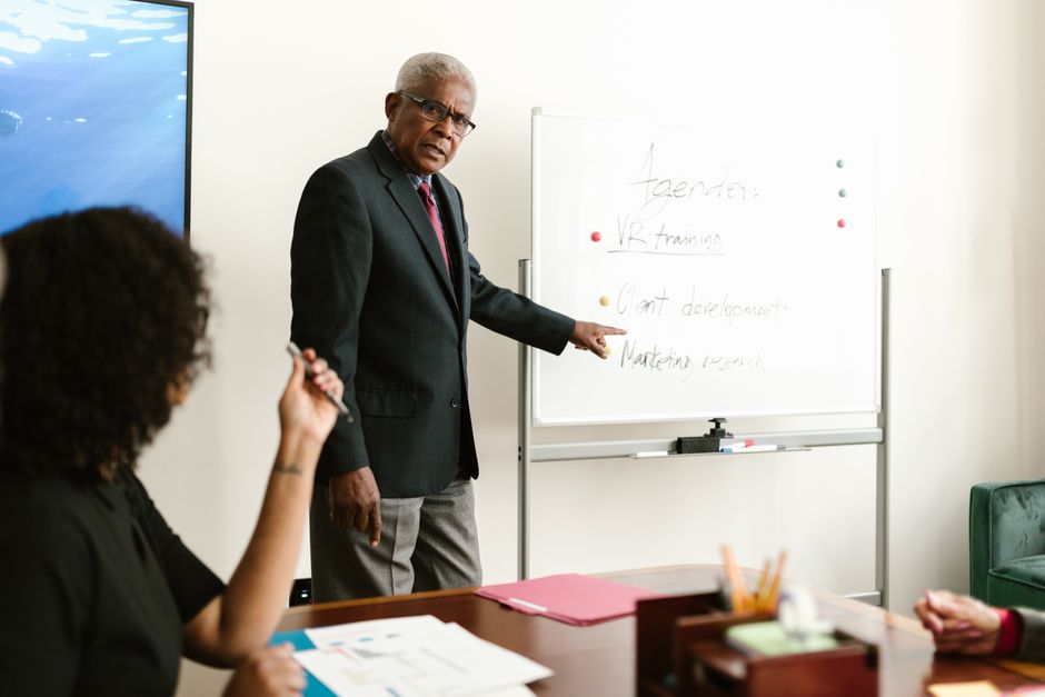 An elderly man in business attire leading a meeting and discussing plans on a whiteboard.