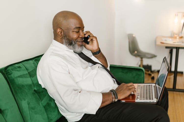 A Man In White Long Sleeves Talking On The Phone While Using His Laptop