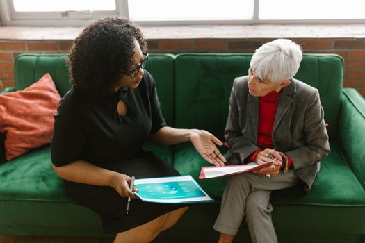Two women in a business meeting discuss documents while sitting on a green sofa.