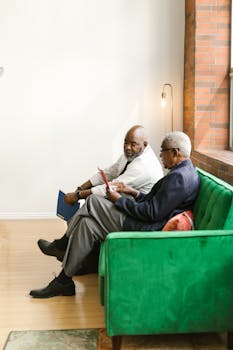 Two businessmen sitting on a green couch engaging in conversation with documents.