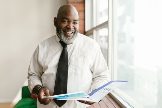 Smiling man with beard and business attire holding papers indoors near a window.