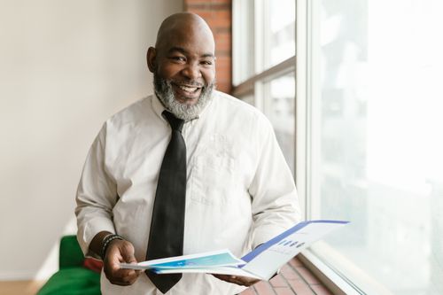 Free Smiling man with beard and business attire holding papers indoors near a window. Stock Photo