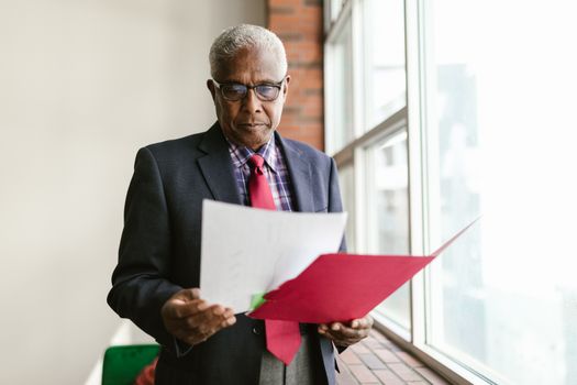 Elderly man in business attire examining papers by a window in an office setting.