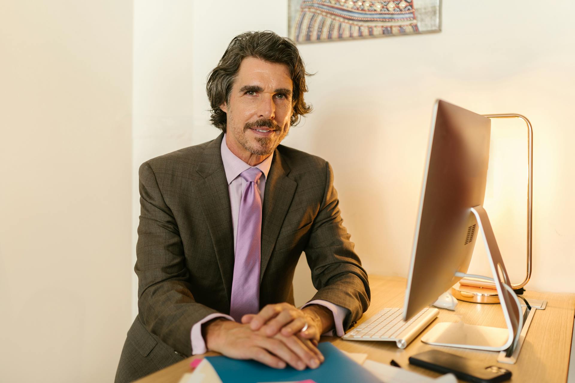 Senior businessman in a gray suit smiles while working at his desk in an office setting.