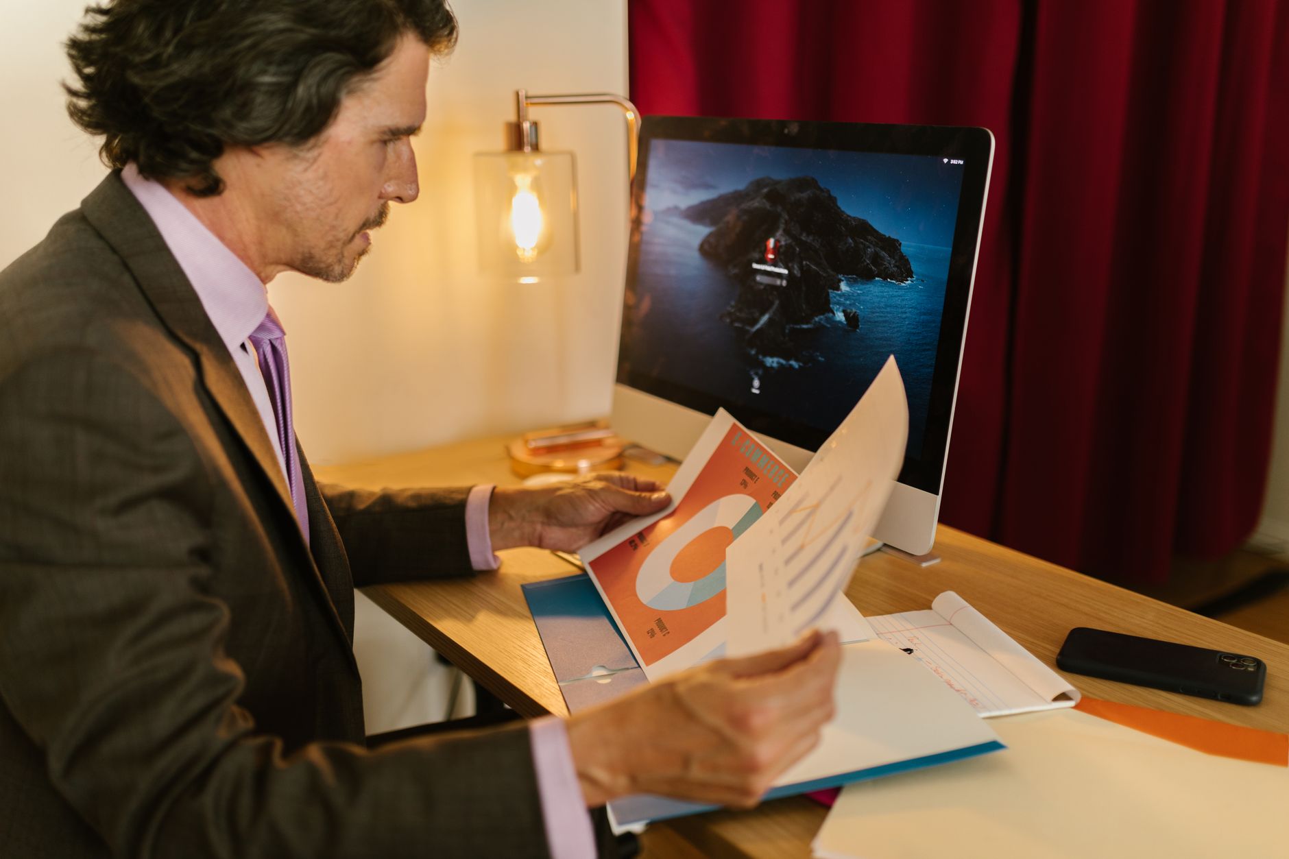 A business professional in a brown suit analyzes printed reports at a desk with a computer.