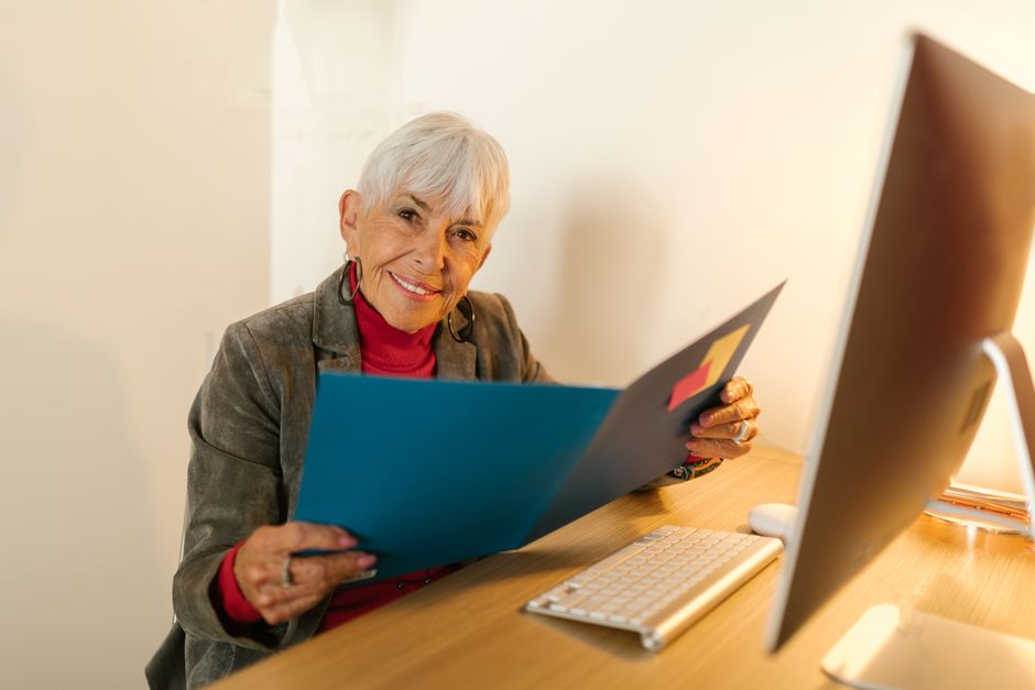 Mujer mayor vestida de negocios sentada en un escritorio, sosteniendo una carpeta y sonriendo cálidamente en un entorno de oficina.