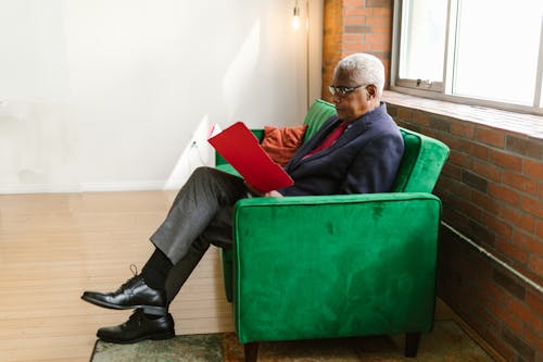 An Elderly Man in Blue Suit Sitting on the Couch while Looking on Red Folder