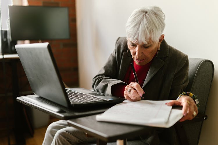 An Elderly Woman Writing On Paper Near The Laptop