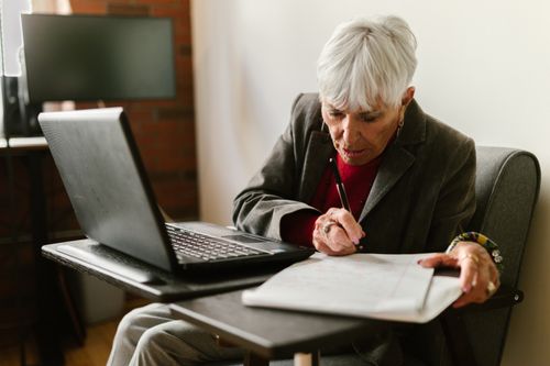 Free Elderly woman in business attire writing on paper beside a laptop in a modern office setting. Stock Photo