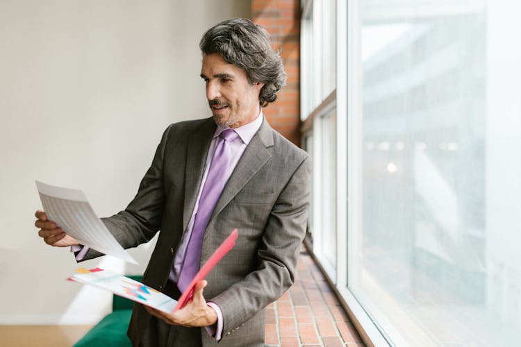 A Man In Gray Suit Looking At The Papers While Standing Near The Window