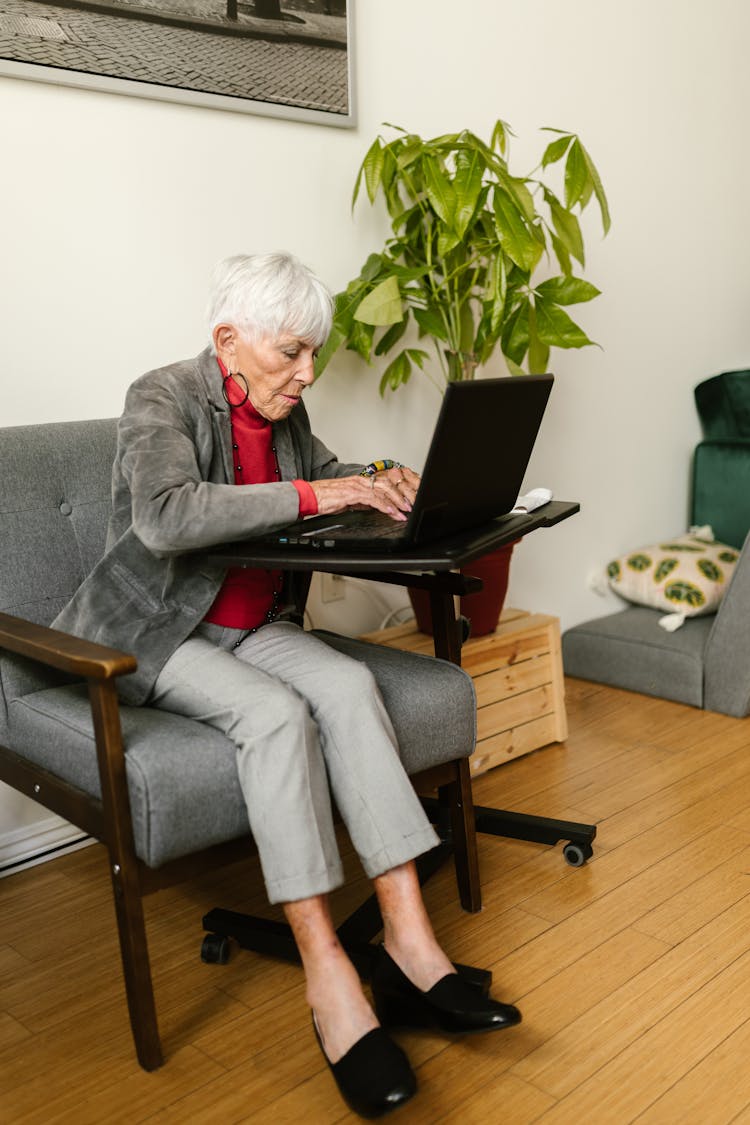 An Elderly Woman In Gray Blazer Sitting On The Chair While Typing On Laptop