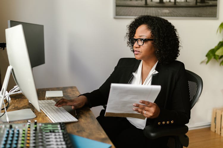 Woman In Black Blazer Holding Papers While Typing On A Keyboard 