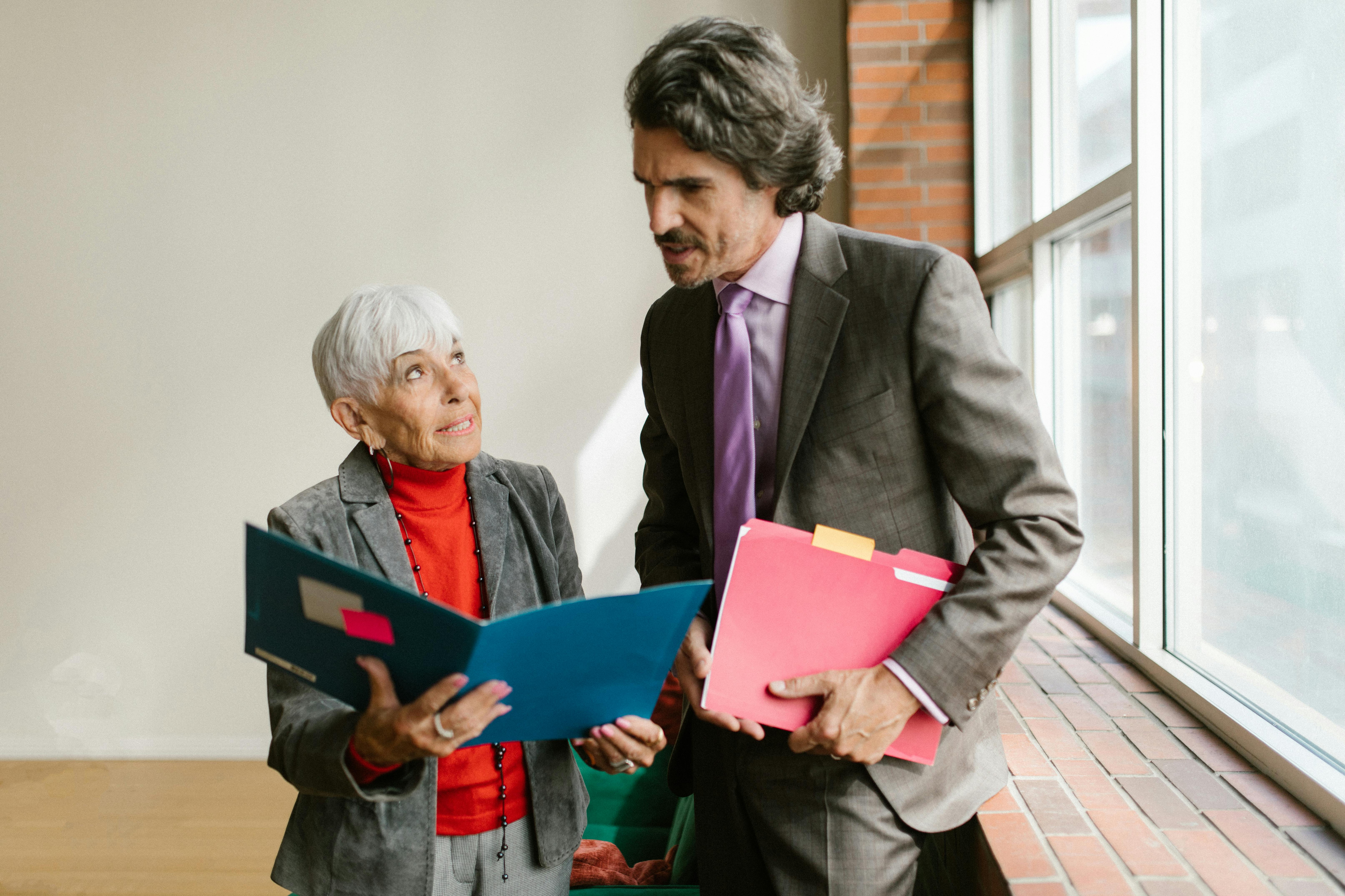 Two senior professionals talking and reviewing business documents near an office window.