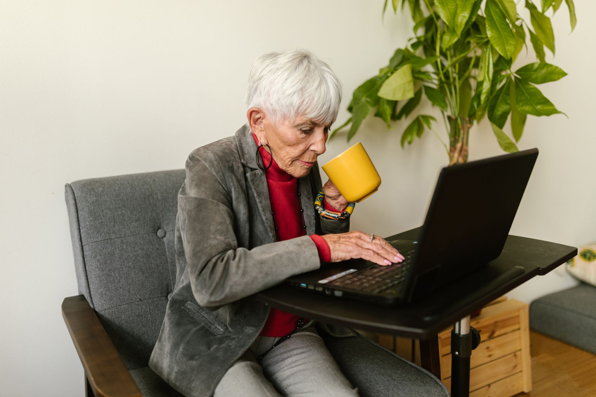 Elderly woman in gray blazer using laptop while drinking coffee, sitting at a home office desk.