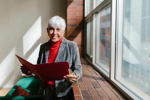 Elderly woman smiling in office attire, holding a folder by a glass window, exuding success and positivity.
