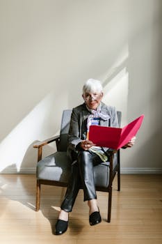 Elderly woman in office attire reading a folder while sitting on a chair in a bright room.