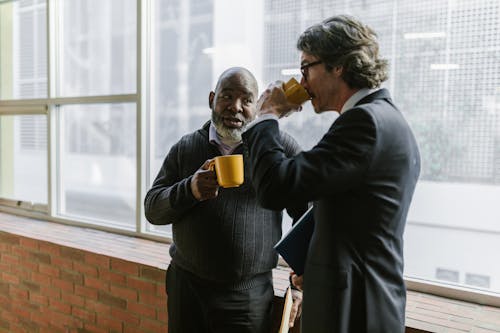 Free Men Leaning on the Windowsill Drinking a Cup of Coffee while Having a Conversation Stock Photo