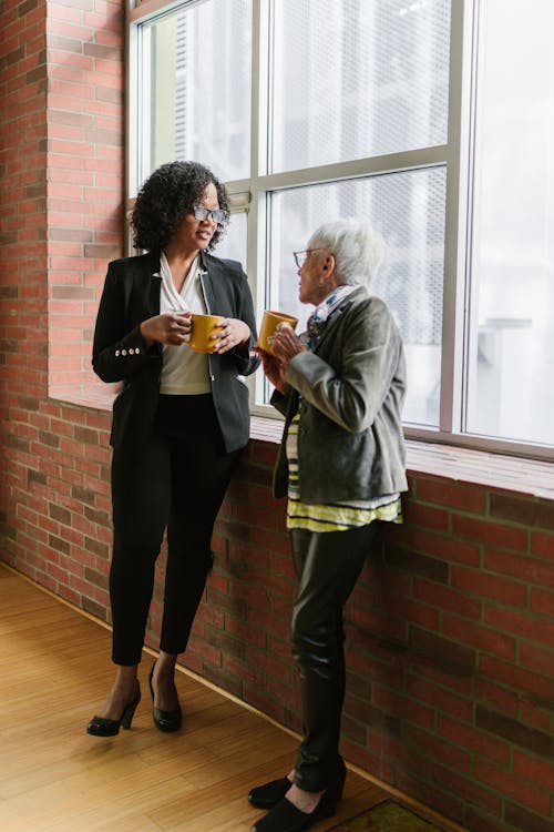 Free A Woman in Black Blazer Talking to the Woman in Gray Blazer while Standing Near the Window Stock Photo