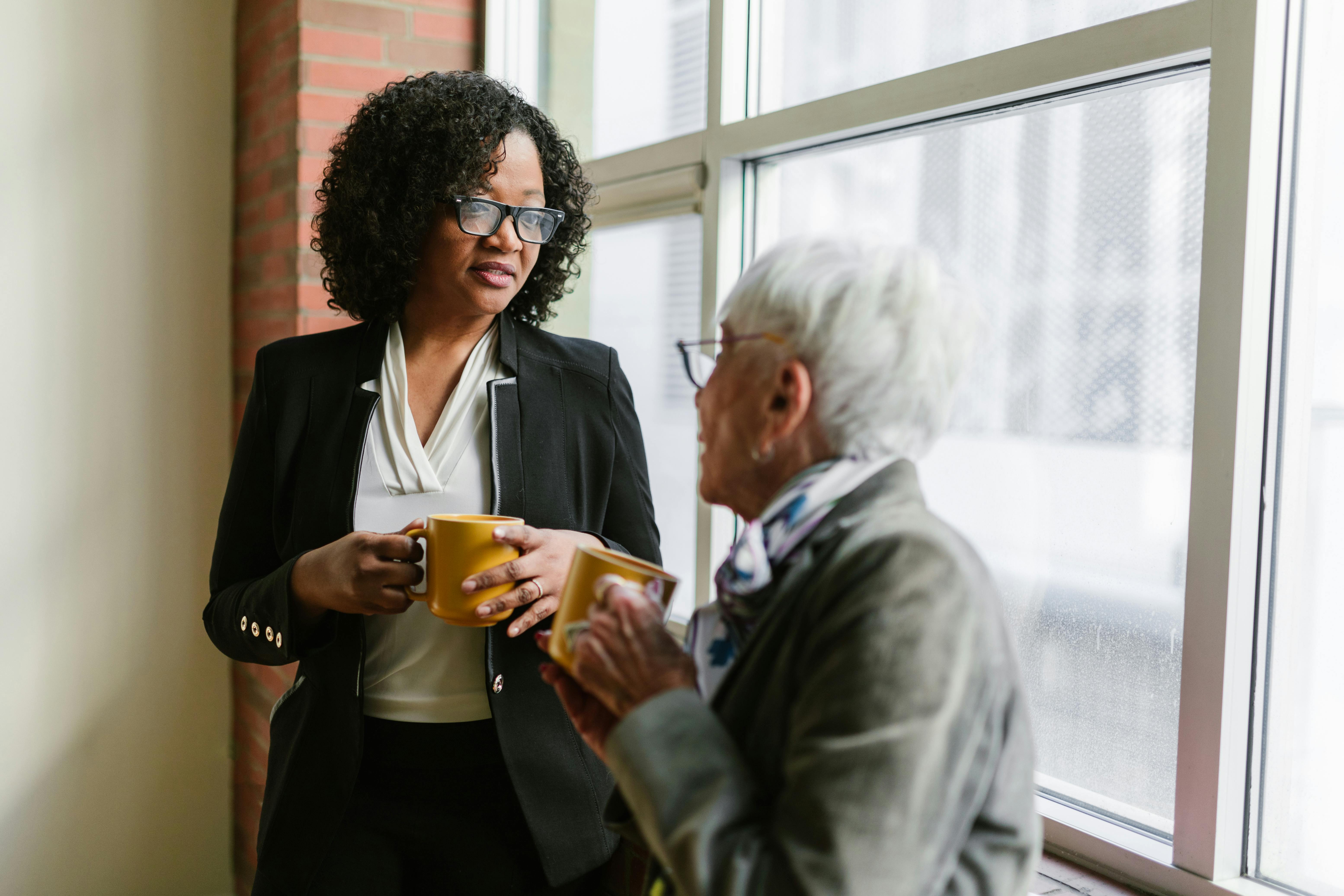 Two women in business attire chatting by a window with coffee mugs in an office setting.