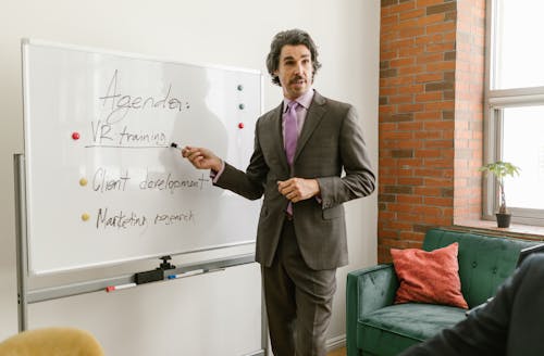 A Man in Gray Suit Talking while Pointing on Whiteboard