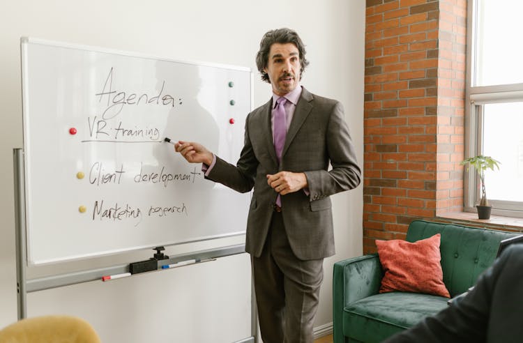 A Man In Gray Suit Talking While Pointing On Whiteboard