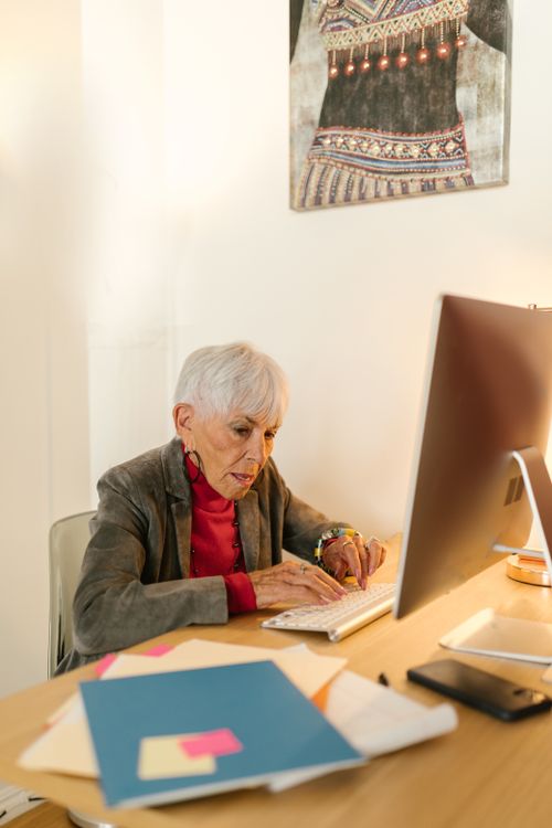 Free An elderly woman in a gray blazer typing on a computer keyboard at her desk in a modern office setting. Stock Photo