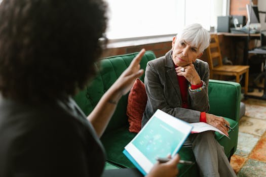 Elderly woman in corporate attire attentively listens during an indoor meeting.