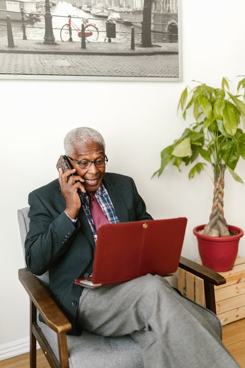 An Elderly Man in Gray Suit Talking on the Phone while Looking at Laptop