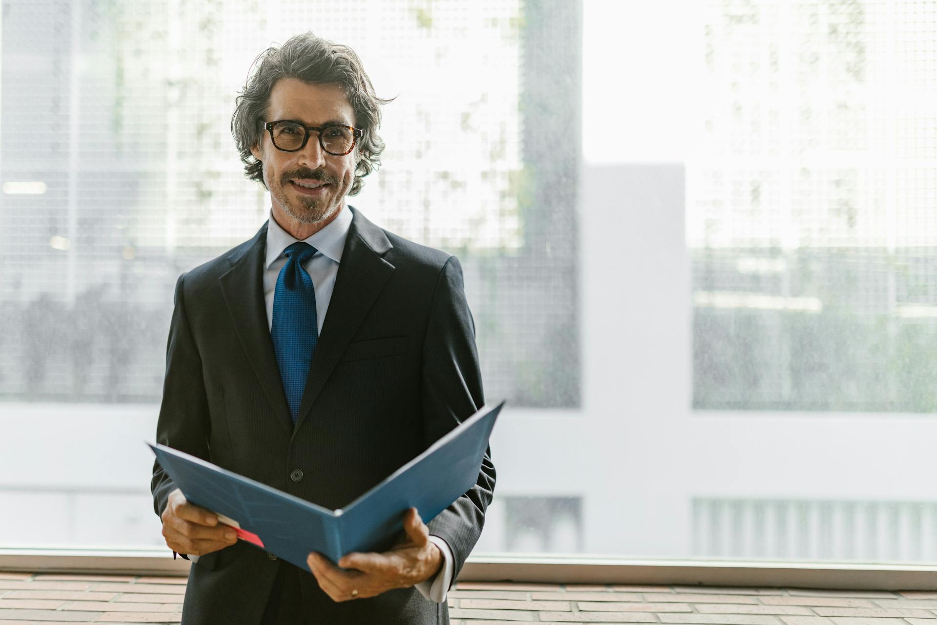 Smiling businessman in a black suit holding a blue portfolio indoors.