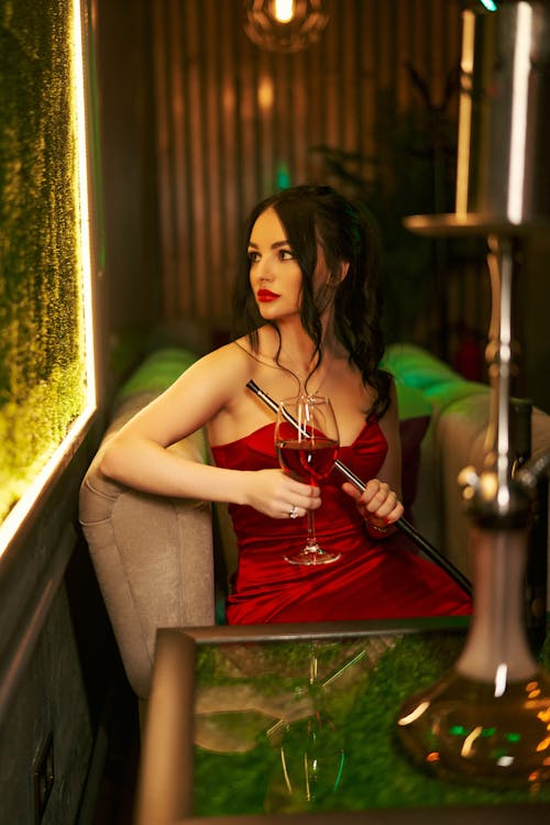 Young alluring female in red dress sitting in comfortable armchair with glass of wine and hookah in restaurant