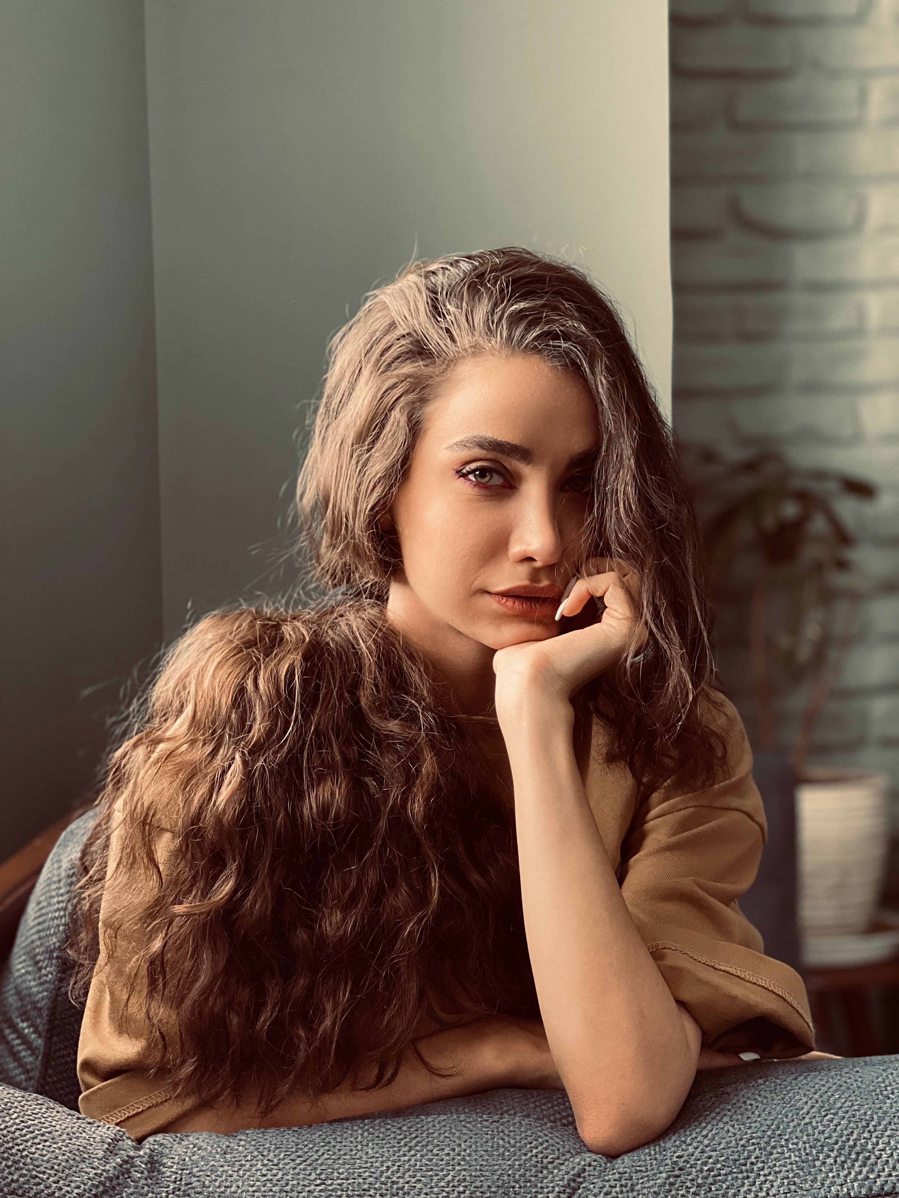 A thoughtful woman with long curly hair posing indoors, captured in a natural light setting.