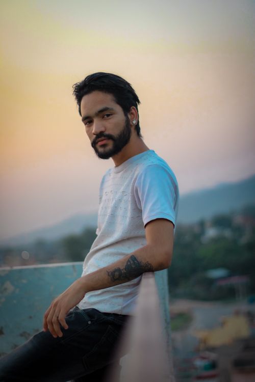 Free A thoughtful man with beard and tattoos leaning on a railing at sunset, wearing a white shirt. Stock Photo