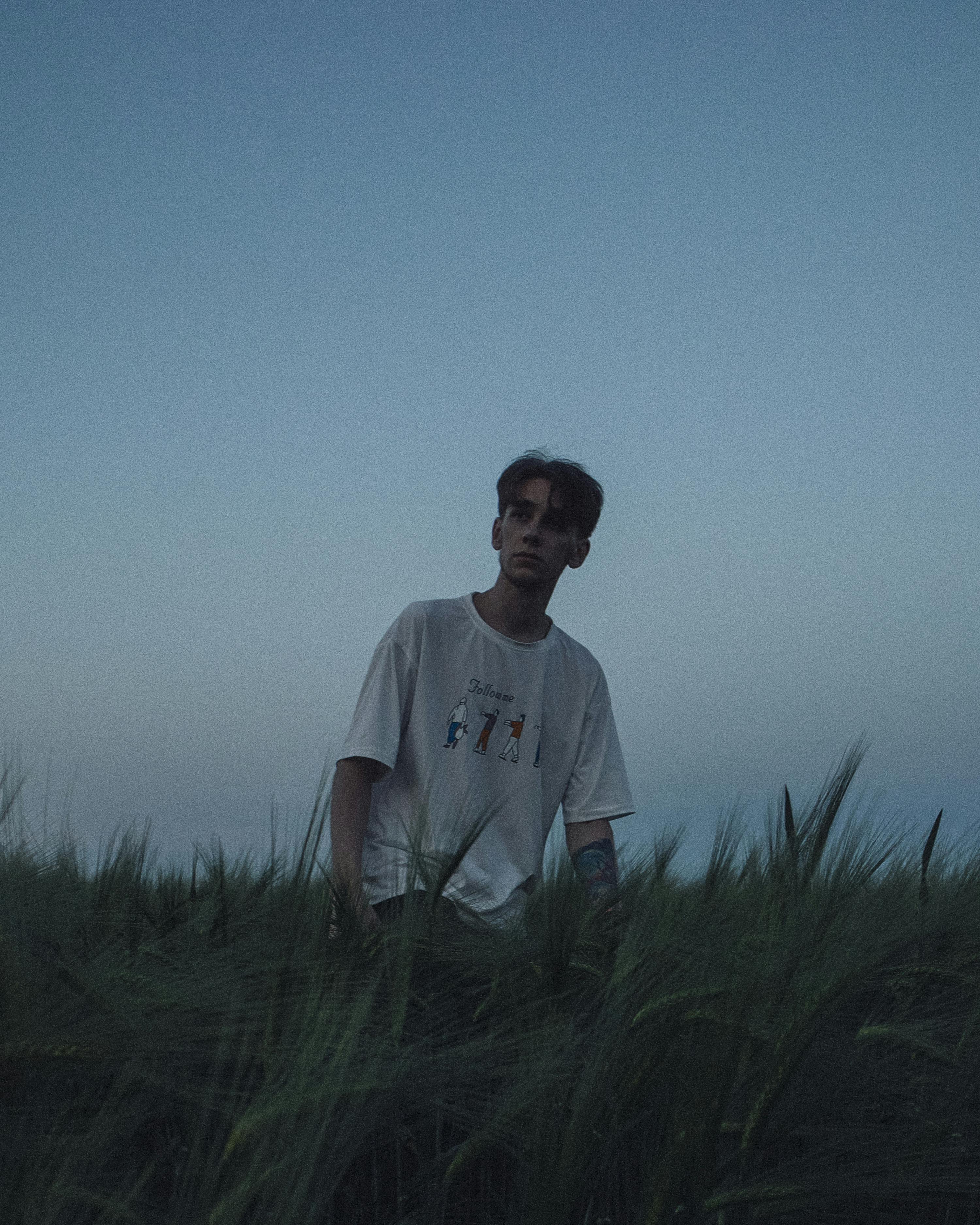 A contemplative young man stands in a field at dusk, wearing a white t-shirt.