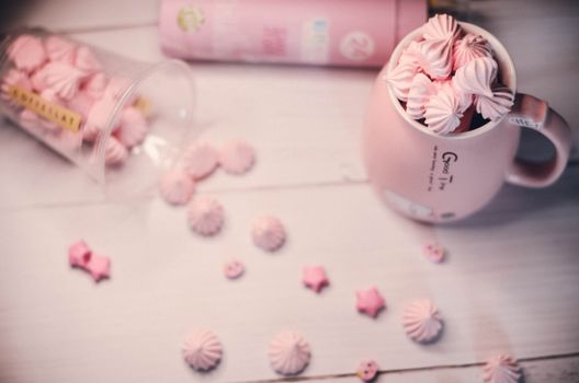 Delightful pink meringues spilling over a ceramic mug on a light wooden surface.