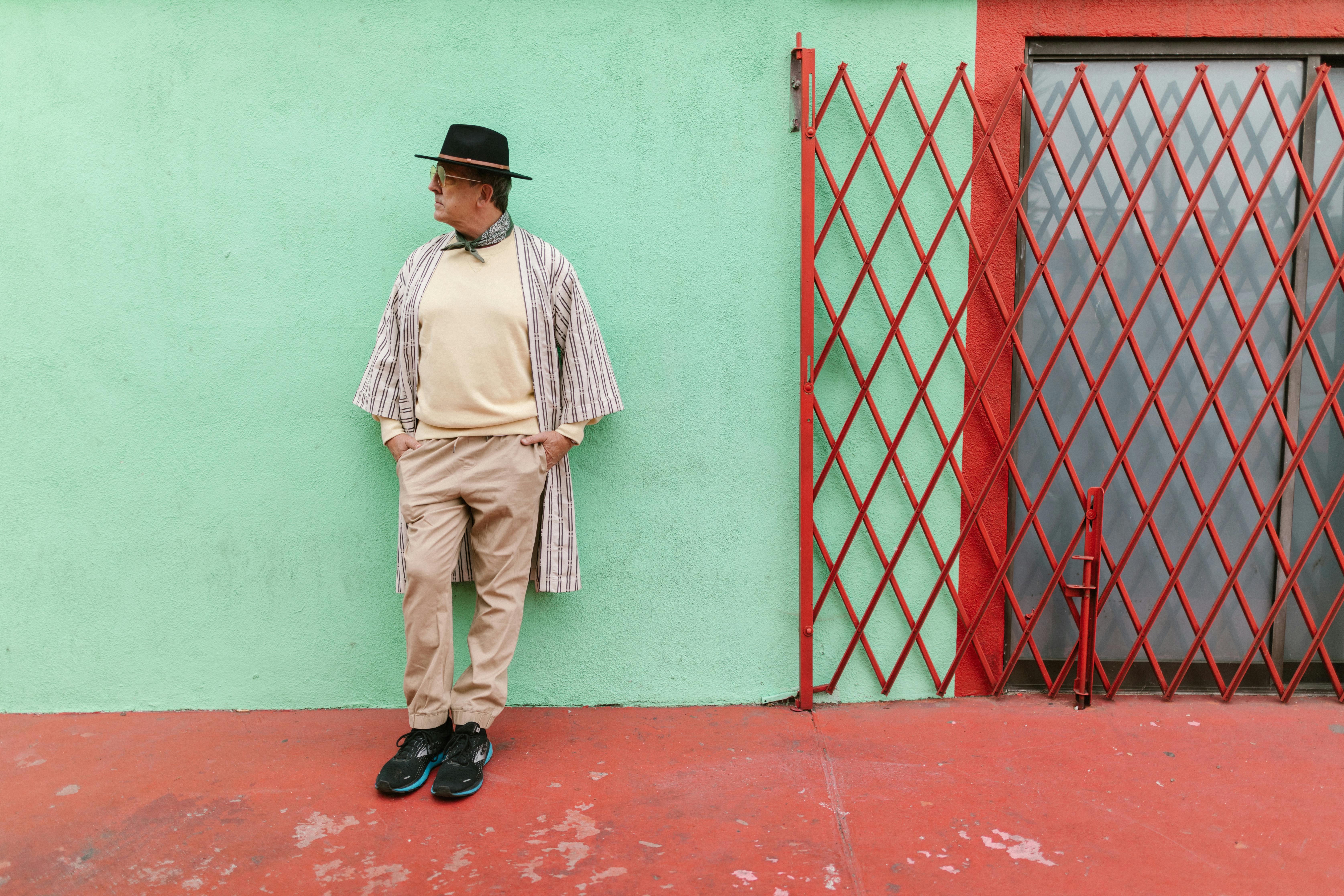 Man in street fashion outfit with fedora hat leaning against a vibrant wall.