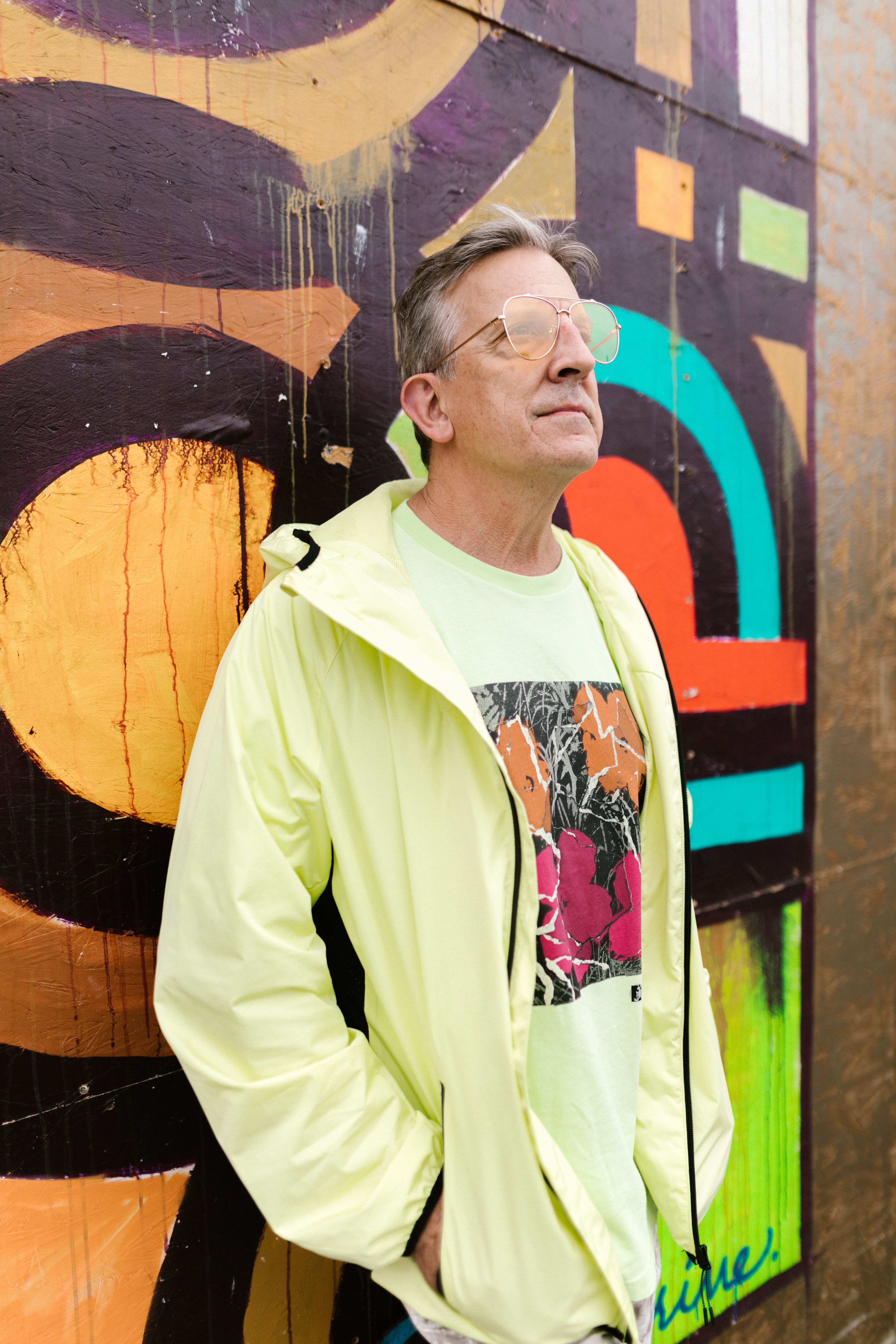 A fashionable man in a green jacket stands against a vibrant graffiti wall.