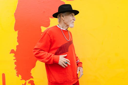 Elderly man in a red sweater and black hat posing against a vibrant wall.
